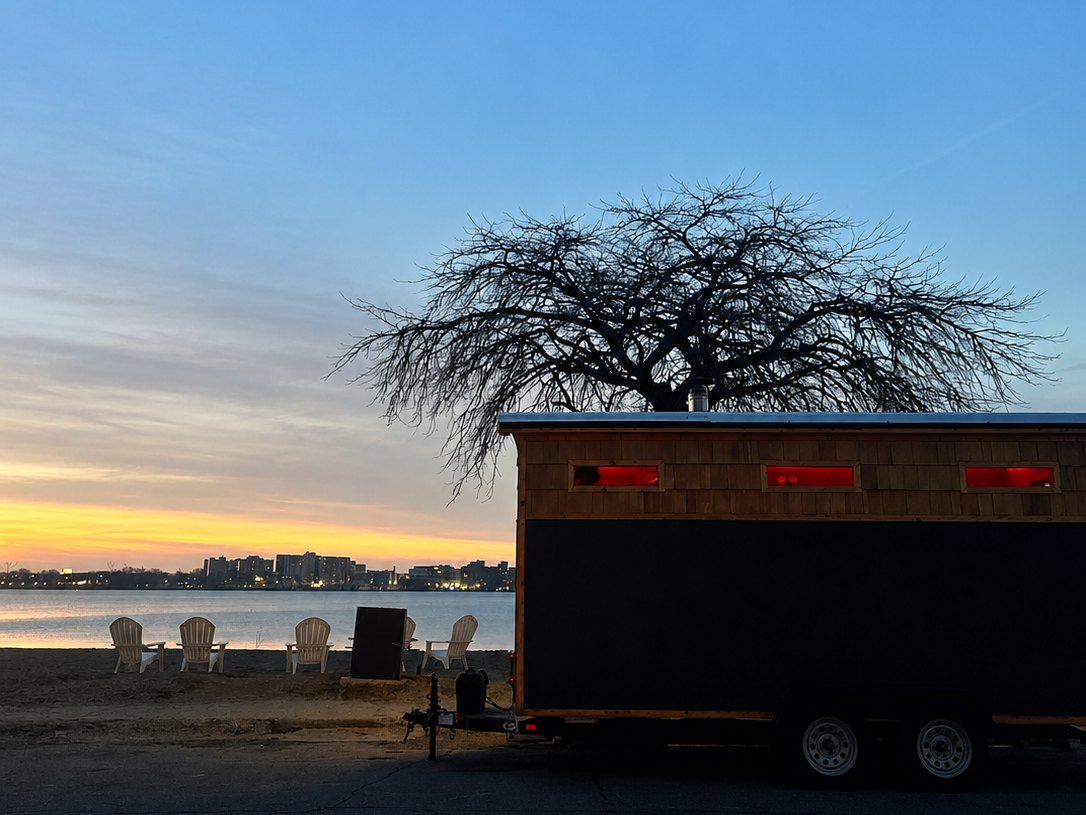 The SOMi cedar sauna on Belle Isle Beach at dusk, chairs on the sand facing the river and skyline
