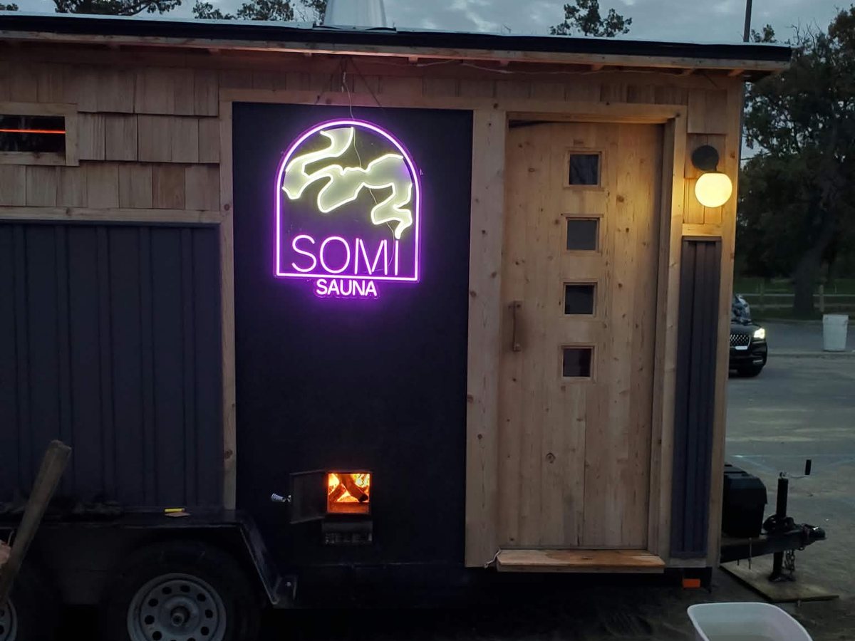 The front of the SOMi sauna at dusk: the neon sign, the cedar door and the wood stove lit